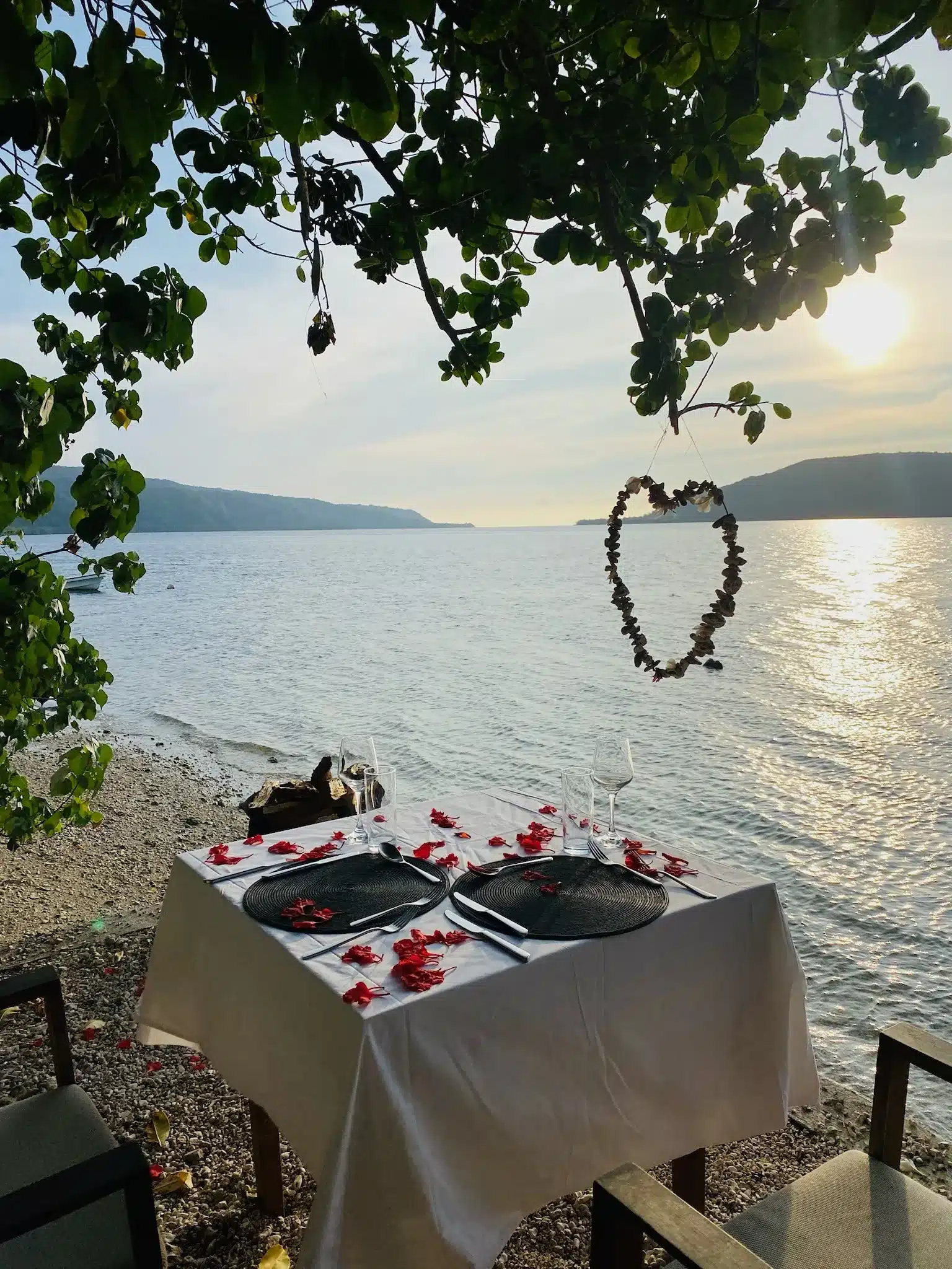 Romantic beachfront dinner table set for two at sunset in Vanuatu, with wine glasses, rose petals and calm water in the background.