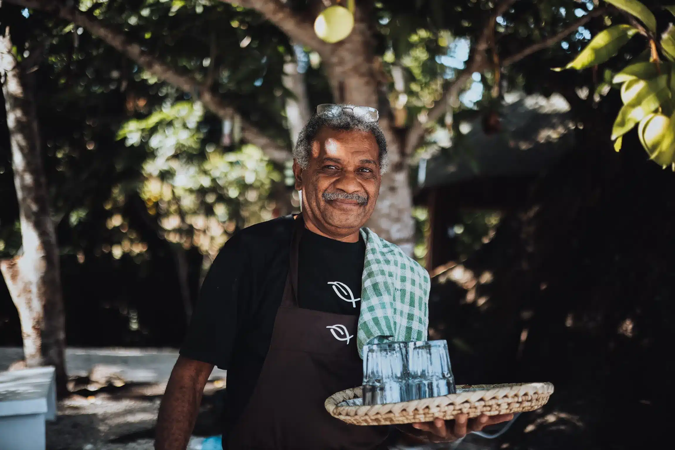 Smiling staff member holding a tray of drinking glasses outdoors at Trees and Fishes in Vanuatu, surrounded by tropical greenery.