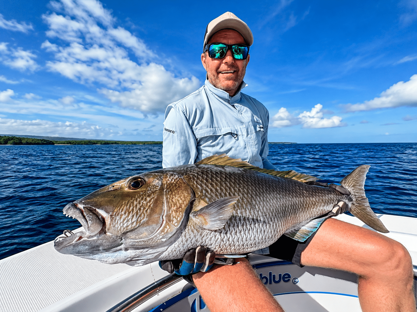 Angler holding a large reef fish on an Ocean Blue Fishing Adventures boat in Vanuatu, with open ocean and blue sky in the background.