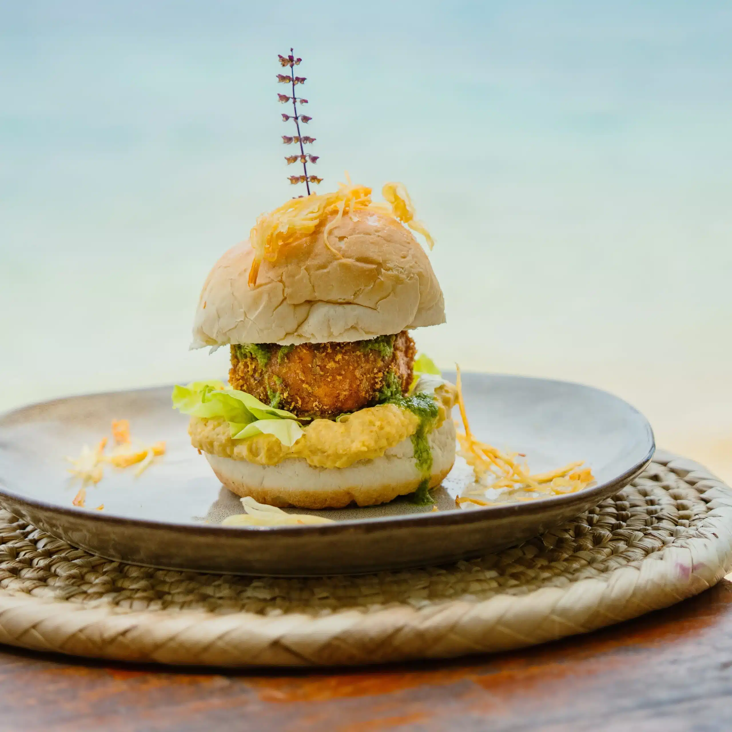 Gourmet burger served on a plate with a crispy crumbed patty, lettuce and green sauce, photographed in a bright tropical dining setting.