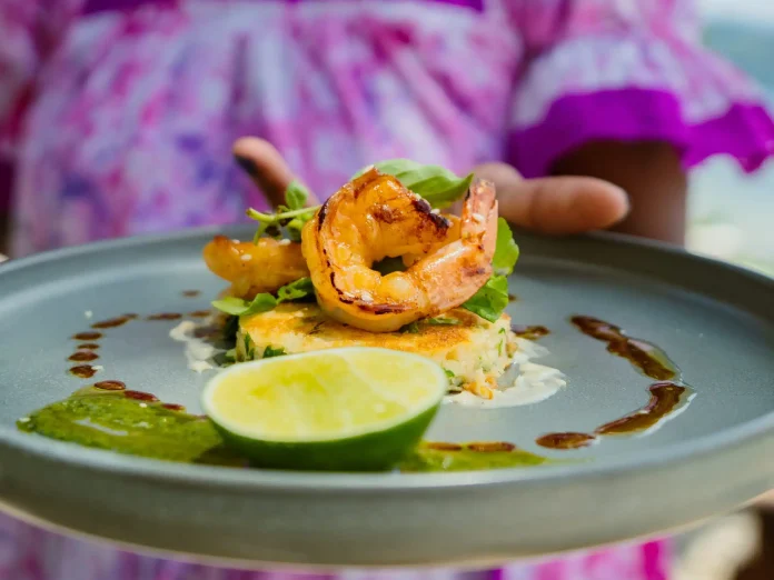 Plated grilled prawn dish served with greens, sauce and a lime wedge, held by a server in a purple dress.